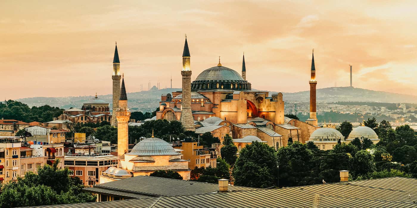 Hagia Sophia at sunset with massive Byzantine dome and four minarets glowing in golden light, surrounding smaller domes and historical buildings, Istanbul cityscape and Bosphorus visible in background under warm peachy sky.