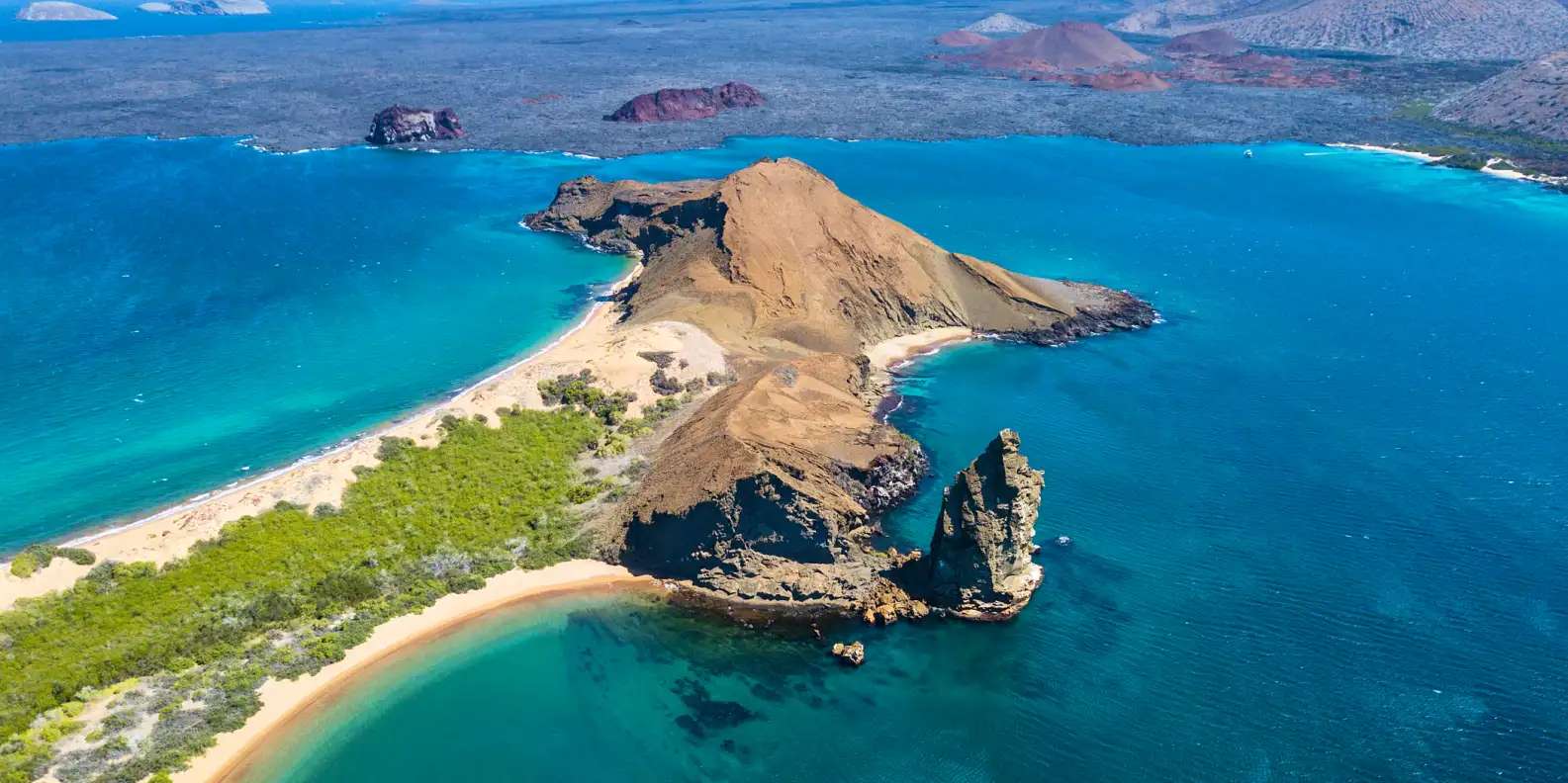 Aerial view of Bartolomé Island in Galápagos showing dramatic volcanic cone rising from turquoise Pacific waters, golden sandy beaches curving around barren landscape with green vegetation, distinctive Pinnacle Rock formation jutting from shoreline.
