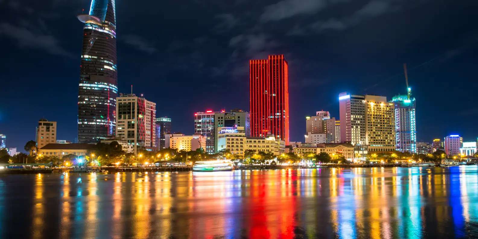 Ho Chi Minh City skyline at night with illuminated skyscrapers including distinctive red-lit tower and colorful building lights reflecting in Saigon River.
