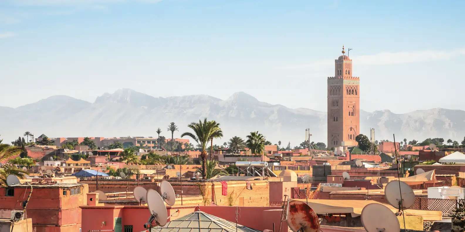 Marrakech cityscape with iconic Koutoubia Mosque minaret towering above terracotta rooftops of medina, palm trees scattered throughout residential buildings, Atlas Mountains visible in hazy distance under pale blue sky.