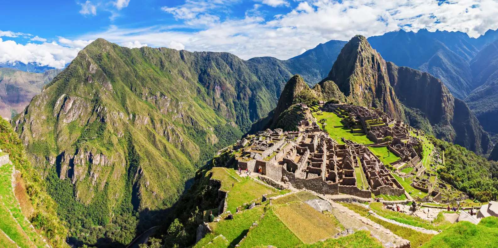 Machu Picchu ancient Inca citadel perched on mountain ridge with iconic Huayna Picchu peak rising behind stone ruins, terraced agricultural platforms with vibrant green grass, steep verdant Andes mountains surrounding site under dramatic cloudy sky.