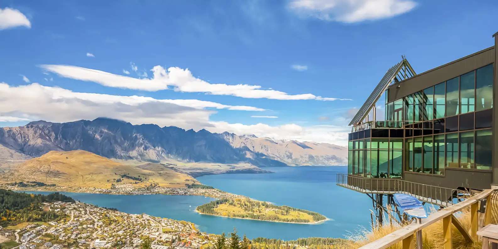 Panoramic view of Lake Wakatipu in Queenstown with modern glass observation deck perched on hillside and the dramatic Remarkables mountain range with snow-capped peaks.
