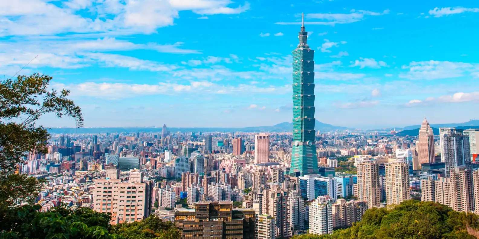 Panoramic view of Taipei cityscape with iconic Taipei 101 skyscraper dominating skyline, turquoise-tinted glass tower rising above dense urban sprawl of modern high-rise buildings, distant mountains visible under bright blue sky with white clouds.