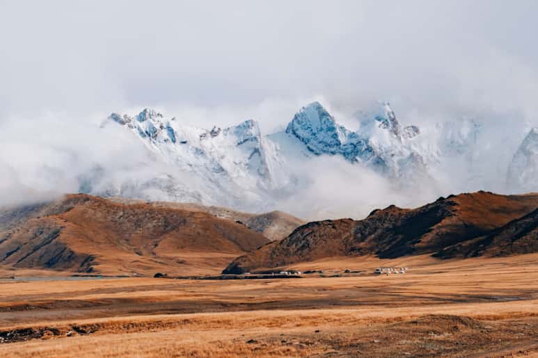 雲霧籠罩着白雪皚皚的山峯,山峯高聳於金棕色的高原之上,前方點綴着零星的居民區和連綿的荒山。